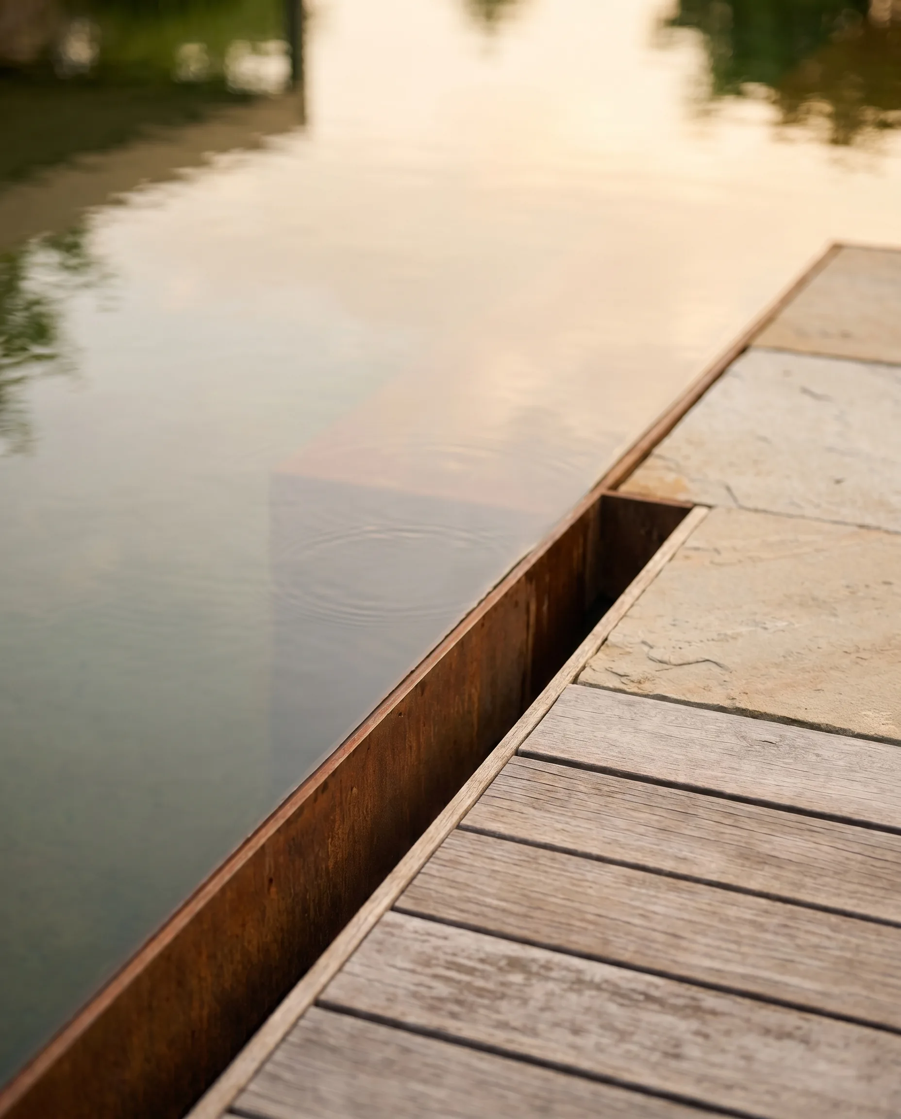 Nahaufnahme einer modernen Gartengestaltung: Cortenstahl-Teichkante trifft auf Holzdeck im warmen Abendlicht.