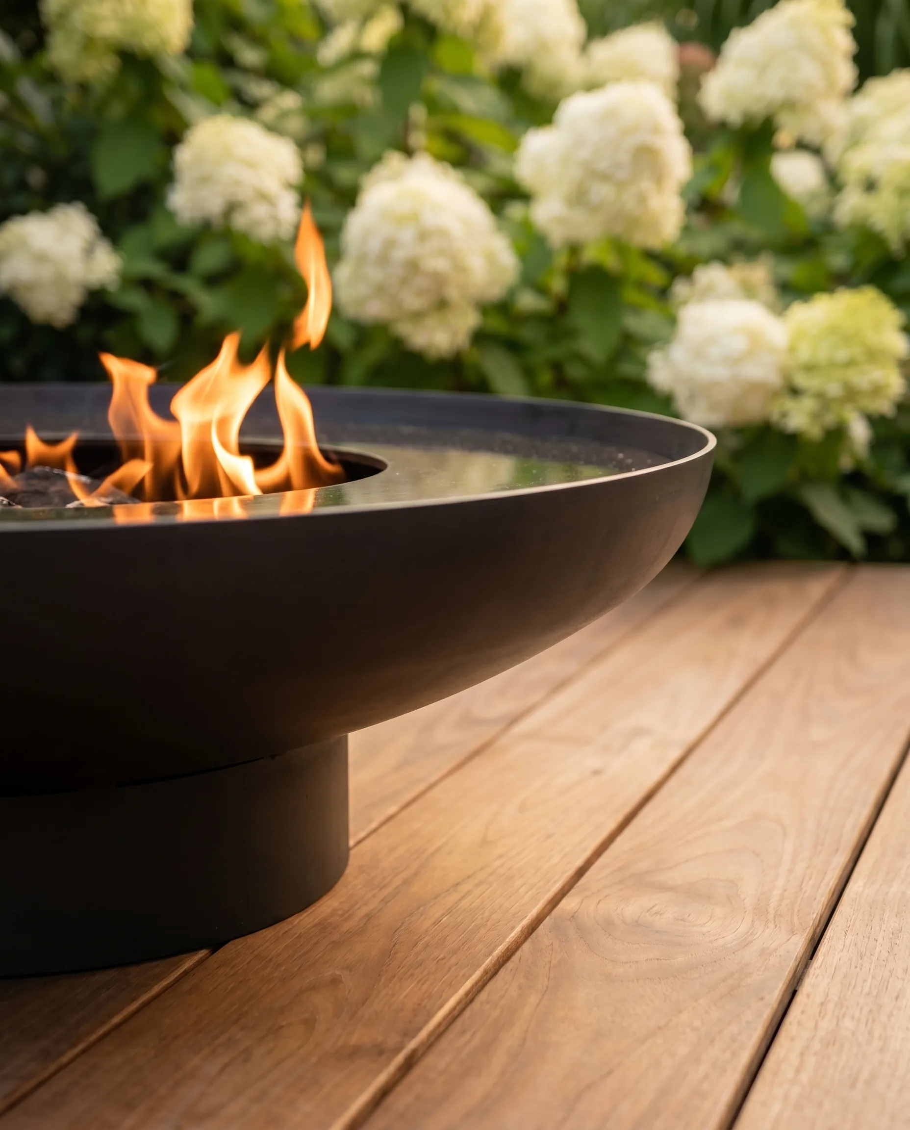 Moderne Gartengestaltung im Detail: Schwarze Stahlschale mit offener Flamme auf Holzdeck, weiße Hortensien sanft unscharf im Hintergrund.